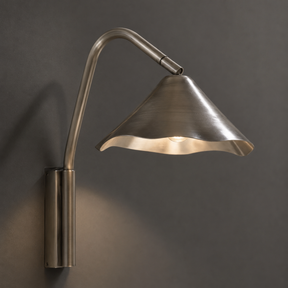 A modern wall-mounted sconce with a brushed metal finish, featuring a curved gooseneck arm and a flared shade that casts a warm light against a dark gray wall.