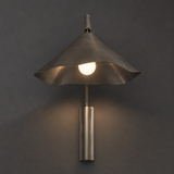 A modern wall-mounted sconce with a flared brushed metal shade and a cylindrical base, shown lit against a dark gray wall.