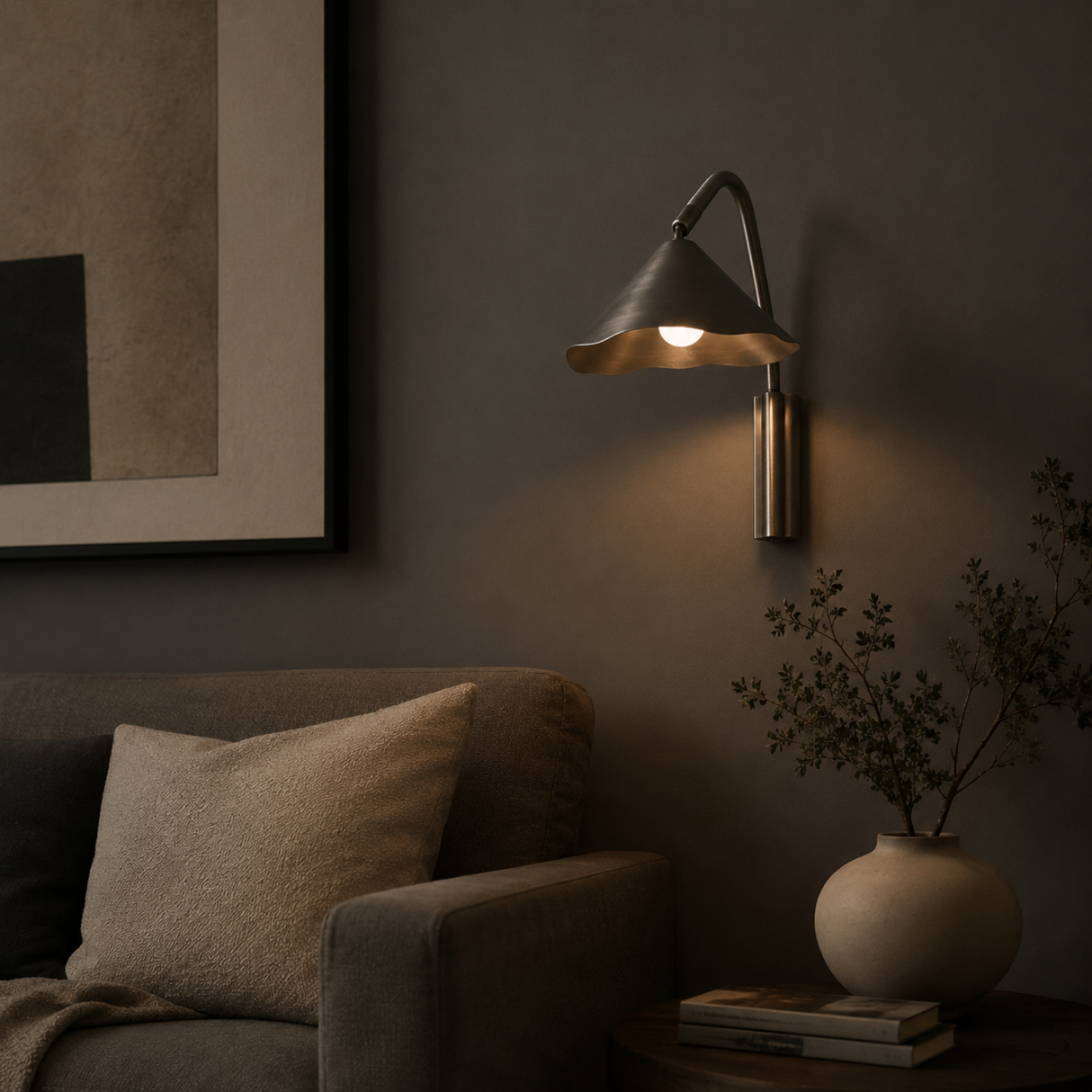 A modern wall-mounted sconce with a conical shade and curved arm, shown lit on a dark gray wall above a sofa in a styled interior setting.