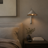 A modern wall-mounted sconce with a conical metallic shade and brushed metal stem, glowing with warm light on a bedroom wall above a bedside table.