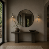 A minimalist interior scene with a round wall mirror centered above a dark wood console table. A large ceramic vase with delicate branches sits on the left side of the table, and a dark bowl is on the right.