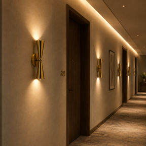 A modern wall sconce with a brushed gold finish mounted on a beige hallway wall, casting a warm light downward. The hallway features dark wood doors, framed wall art, and recessed ceiling lighting.