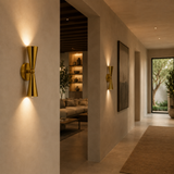 A modern wall-mounted sconce in brushed gold metal with a sculptural hourglass shape, shown lit in a warm, contemporary hallway setting.