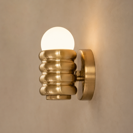 A modern wall-mounted sconce with a ribbed metallic gold shade and a glowing frosted globe bulb, mounted on a textured beige wall.