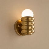A modern wall-mounted light fixture with a ribbed metallic gold shade and a glowing white globe bulb, mounted on a warm beige wall.