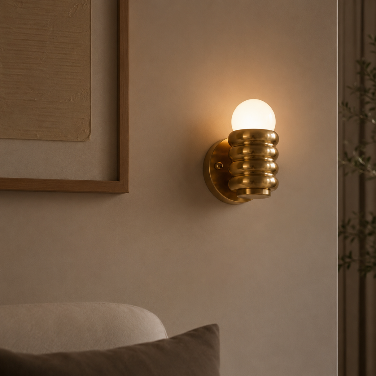 A modern wall sconce with a ribbed metallic gold body and a glowing warm white globe bulb, mounted on a beige wall in a styled interior setting.