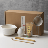 Tea-making set with a brown box, ceramic bowl, whisk, scoop, and tea leaves on a gray background.