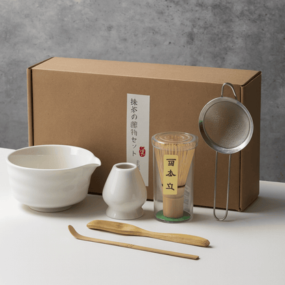 Tea-making set with a brown box, ceramic bowl, whisk, scoop, and tea leaves on a gray background.