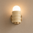 A wall-mounted light fixture with a glowing frosted globe bulb, a ribbed cream glass shade, and a round gold metal base against a warm beige wall.