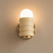 A wall-mounted light fixture with a glowing frosted globe bulb, a ribbed cream glass shade, and a round gold metal base against a warm beige wall.
