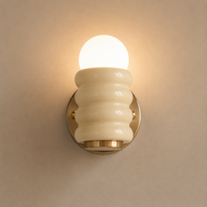 A wall-mounted light fixture with a glowing frosted globe bulb, a ribbed cream glass shade, and a round gold metal base against a warm beige wall.