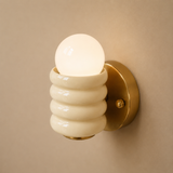 A modern wall-mounted sconce with a glowing frosted glass globe bulb, a ribbed cream-colored shade, and a brushed brass circular wall mount against a warm beige wall.