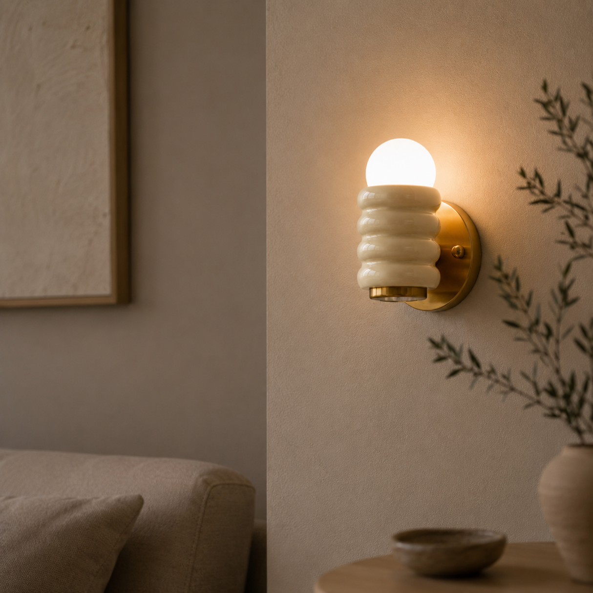 A modern wall sconce light fixture with a ribbed white glass shade and a gold brass backplate, glowing with a warm light on a beige textured wall in a styled interior setting.