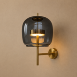 A modern wall sconce light fixture with a large, rounded translucent dark gray glass shade and a brushed brass metal arm and wall mount, shown illuminated against a warm beige wall.