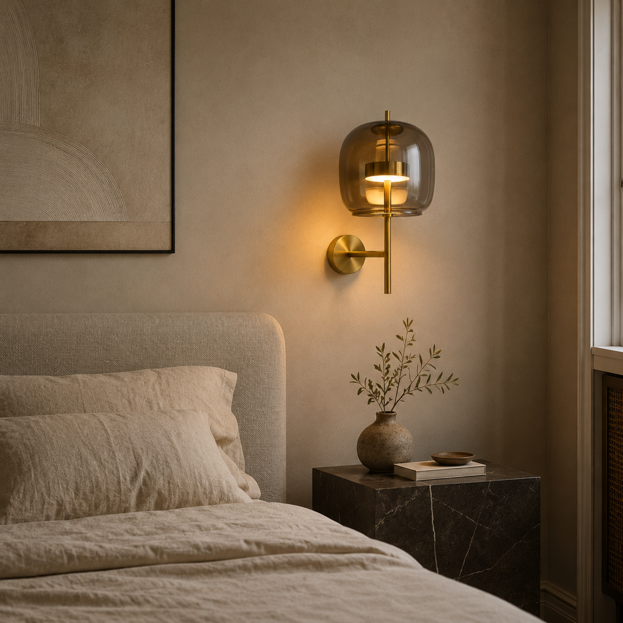 A modern wall-mounted sconce with a warm brass finish and a translucent amber-tinted glass dome, illuminated in a cozy bedroom setting next to a bed.