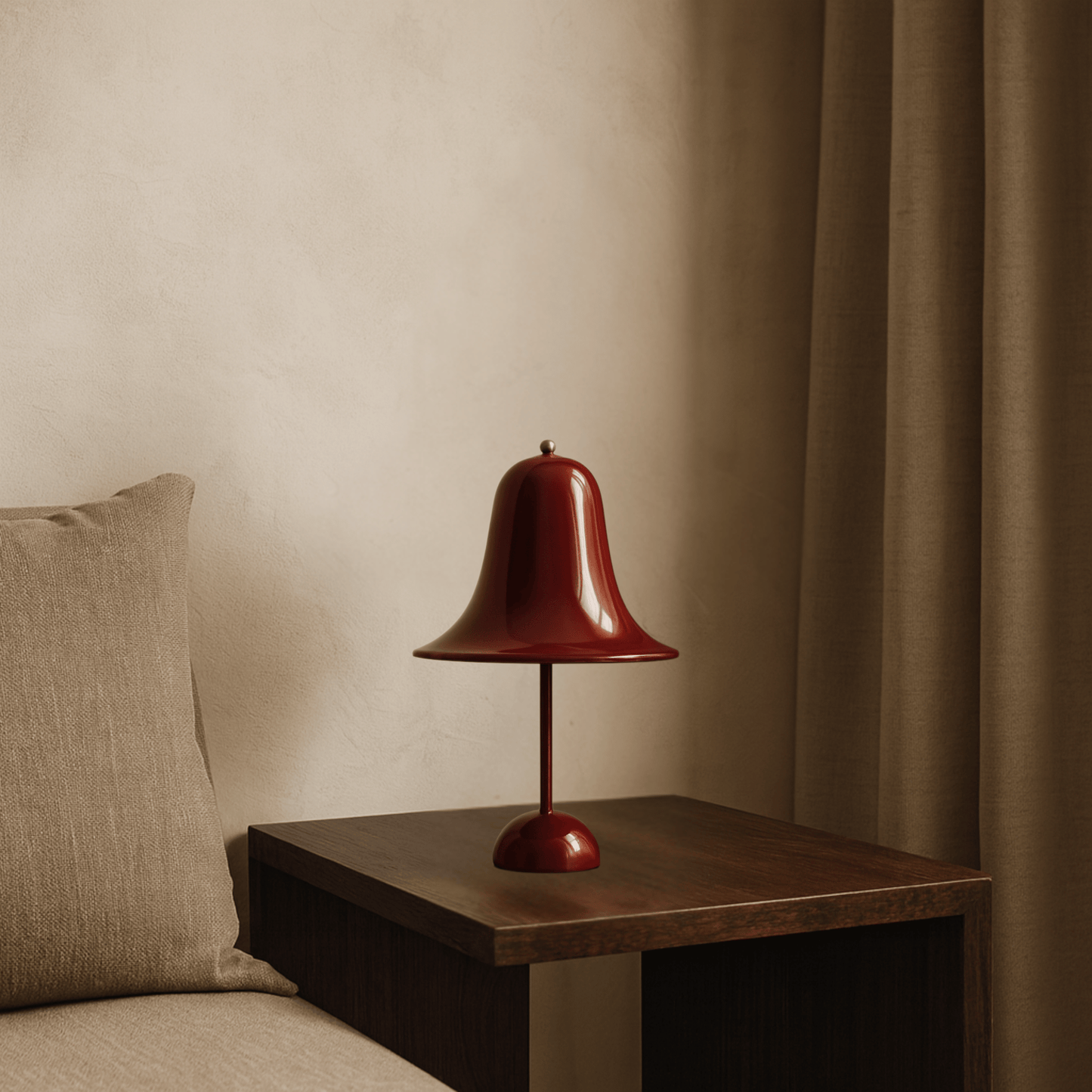 Red bell-shaped table lamp on a wooden side table next to a beige pillow against a beige wall.