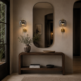 A modern interior scene with a tall rounded-rectangle wall mirror centered above a dark wood console table. Two matching wall sconces with smoky glass shades and gold bases are mounted on either side of the mirror, casting warm light.