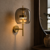 A modern wall-mounted sconce with a clear glass dome shade and brushed brass hardware, lit warmly against a textured beige wall.