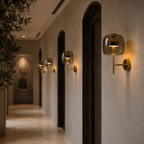 A modern wall-mounted sconce with a smoked amber glass dome and a gold-finished arm and base, illuminated in a hallway with beige walls and dark arched doorframes.