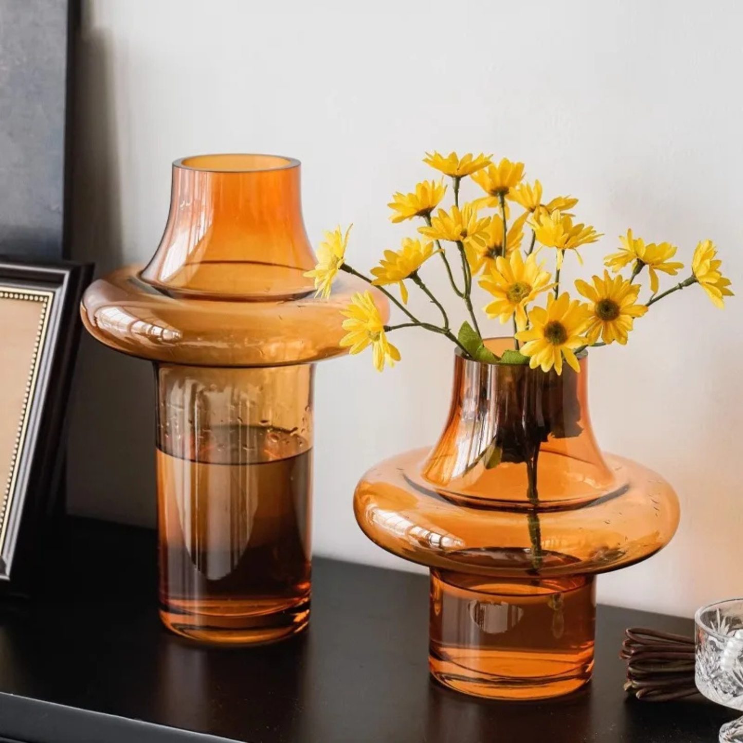 Two amber glass vases placed on a shelf, one holding bright yellow flowers and the other filled with water, next to framed decor.
