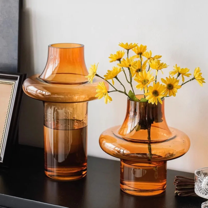 Two amber glass vases placed on a shelf, one holding bright yellow flowers and the other filled with water, next to framed decor.