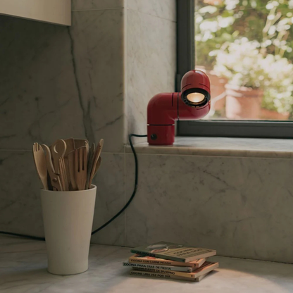 A red desk lamp in an elbow shape placed near a window, illuminating a countertop with wooden utensils and cookbooks.