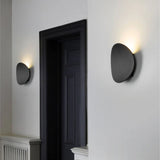 A black circular wall lamp glowing on a white wall near a dark doorway in a contemporary interior.