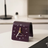 A purple leather desk clock with golden hands on a white desk beside a lamp and minimal decor.