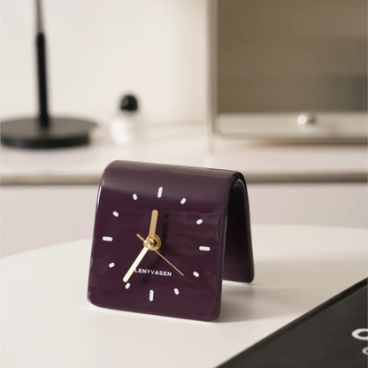 A purple leather desk clock with golden hands on a white desk beside a lamp and minimal decor.