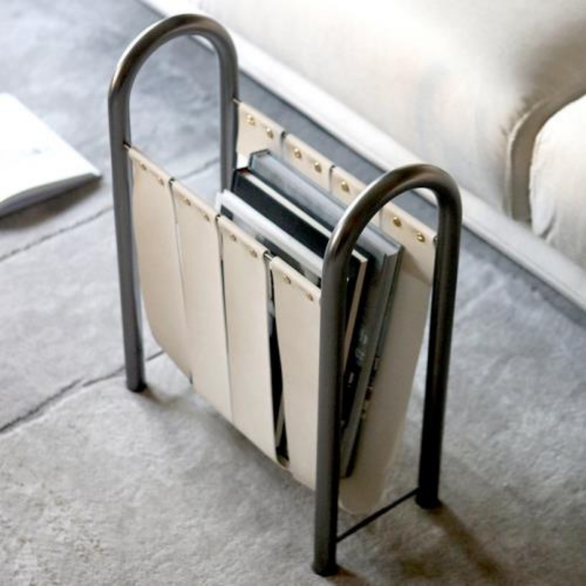 A tall, white leather and metal magazine holder standing upright on a soft gray carpet next to a sofa.