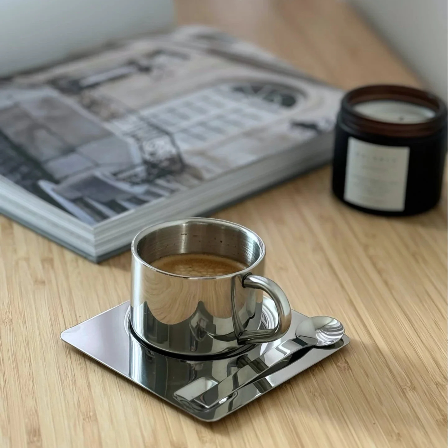 Top view of a stainless steel coffee cup filled with foam coffee on a reflective square saucer with a metal spoon.
