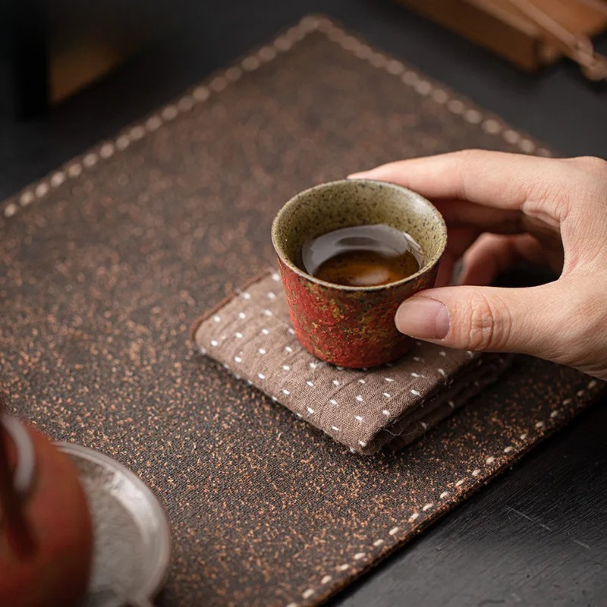 A hand holding a red ceramic tea cup filled with tea, resting on a folded cloth on a tea table.