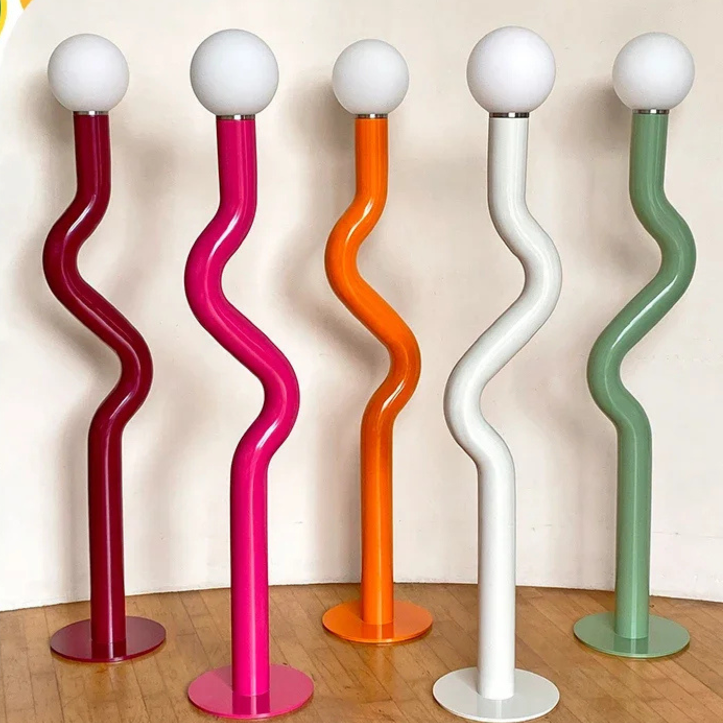 Four vibrant zigzag-shaped floor lamps in red, pink, orange, and white arranged beside a wall.
