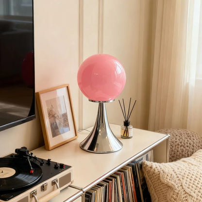 A pink glass globe table lamp with a chrome base positioned on a white surface near a window with warm sunlight.