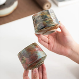 Close-up of two multicolor ceramic cups on a desk, one filled with coffee and being stirred with a small gold spoon.