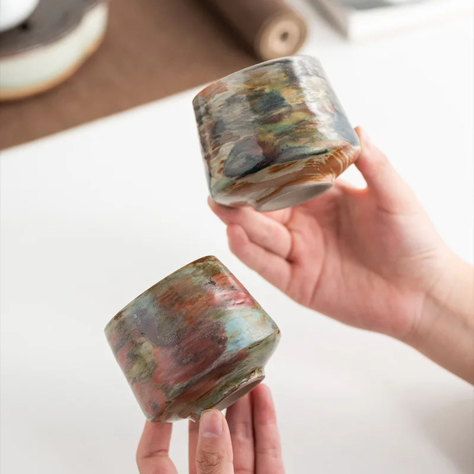 Close-up of two multicolor ceramic cups on a desk, one filled with coffee and being stirred with a small gold spoon.