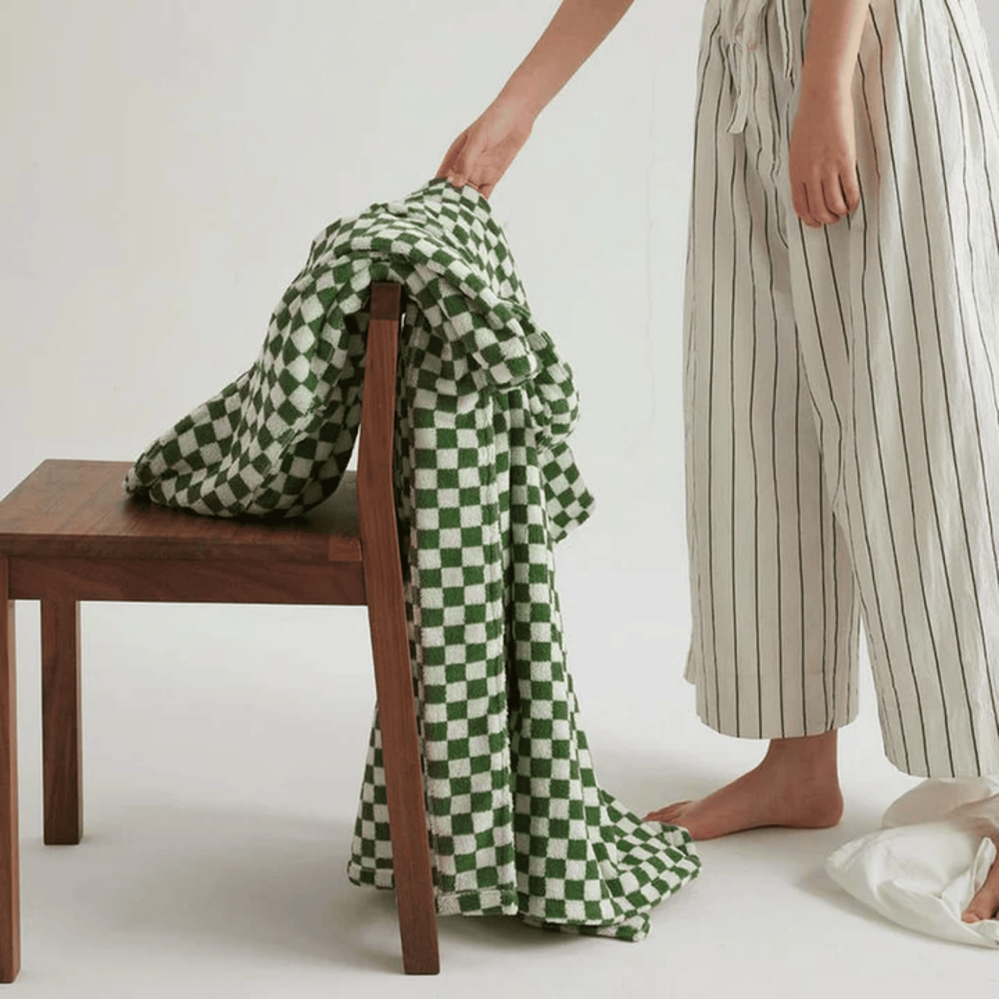 Person holding a green and white checkered blanket next to a wooden chair on a plain background