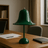 Green table lamp on a wooden desk with a laptop and notebook in a room setting.