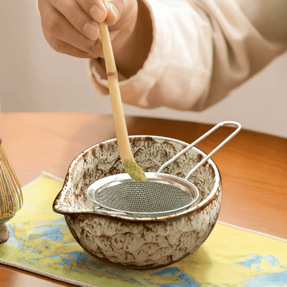 Person using a bamboo whisk to stir tea in a ceramic bowl on a wooden table.
