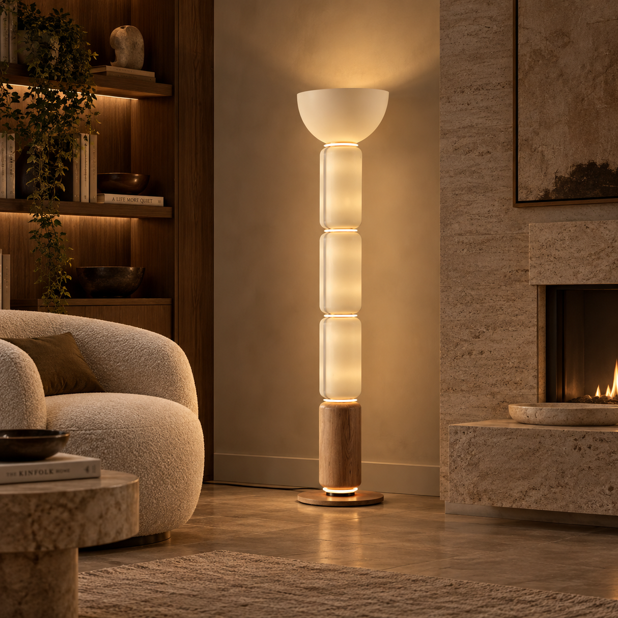 A modern floor lamp with stacked frosted glass cylindrical shades and a warm glowing light, positioned in a cozy, warmly lit living room next to a textured armchair and fireplace.
