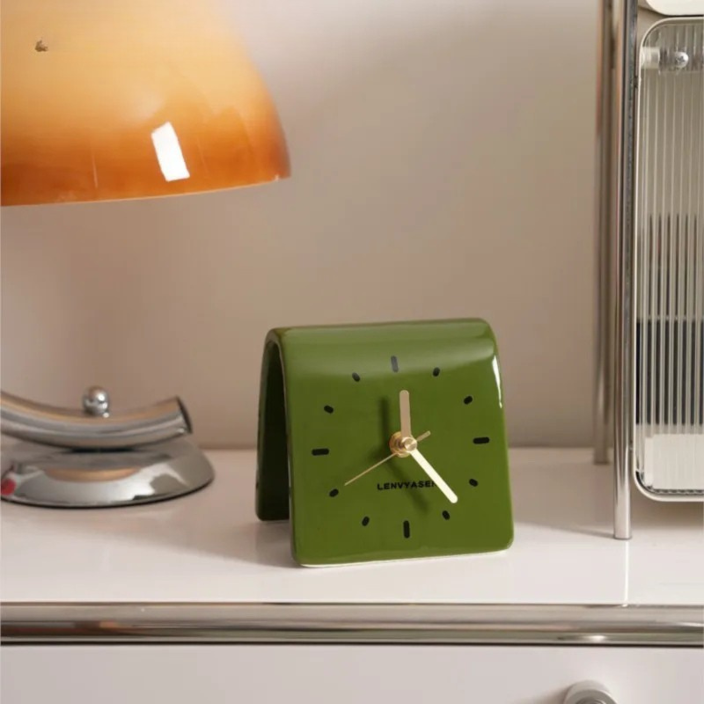 A green leather desk clock with golden hands placed on a table next to an orange lamp.