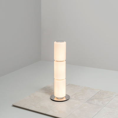 Tall cylindrical floor lamp with ribbed white glass standing on a tiled surface in a minimalist corner.