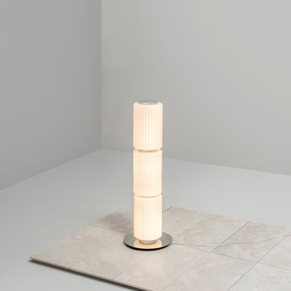 Tall cylindrical floor lamp with ribbed white glass standing on a tiled surface in a minimalist corner.
