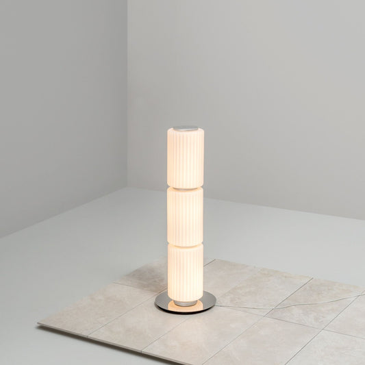 Tall cylindrical floor lamp with ribbed white glass standing on a tiled surface in a minimalist corner.