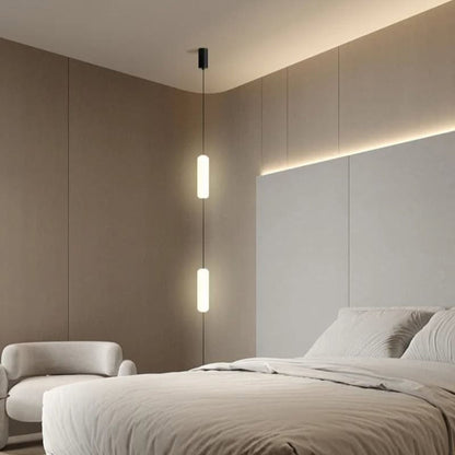 A vertical tube pendant light hanging in a soft, neutral-colored bedroom, illuminating the area near a large window.