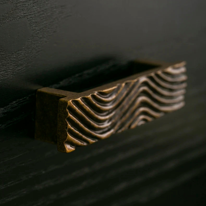 A close-up of a single textured brass handle installed on a dark wood drawer.