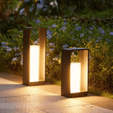Two outdoor lamps on a pathway with greenery and flowers in the background