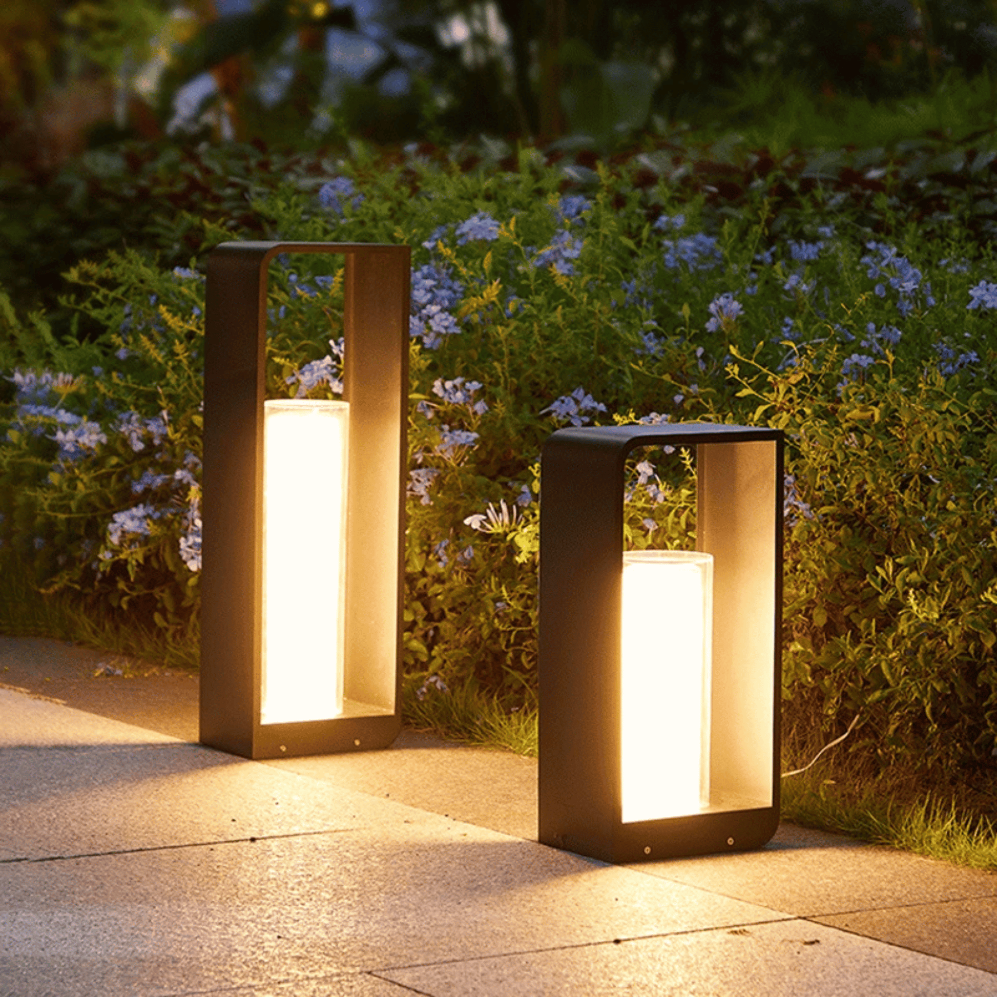 Two outdoor lamps on a pathway with greenery and flowers in the background