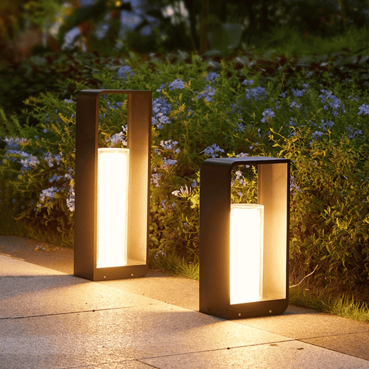 Two outdoor lamps on a pathway with greenery and flowers in the background