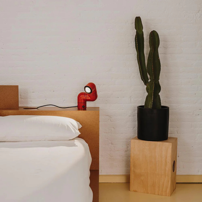 A red elbow-shaped desk lamp next to a bed, casting light on a wooden nightstand and a potted plant.